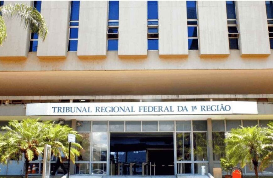 Fachada do prédio do Tribunal Regional Fachada do TRF1 com letreiro institucional, arquitetura moderna de concreto e vidro, céu azul ao fundo e bandeiras do Brasil, Judiciário e TRF1 hasteadas na entrada.