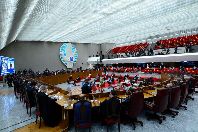 A imagem mostra o plenário do Superior Tribunal de Justiça (STJ) do Brasil durante uma sessão solene. O ambiente é amplo e moderno, com teto translúcido, iluminação clara e assentos vermelhos na galeria superior, onde estão jornalistas e convidados. Os ministros estão sentados em cadeiras de encosto alto em uma mesa circular de madeira, enquanto outras autoridades e participantes ocupam cadeiras no centro do plenário. Há um grande painel decorativo azul com o símbolo do STJ na parede ao fundo e uma tela exibindo pessoas participando remotamente.