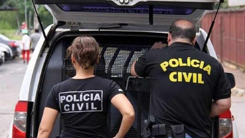 Dois agentes da Polícia Civil, um homem e uma mulher, estão de costas próximos a uma viatura com o porta-malas aberto durante uma ação.