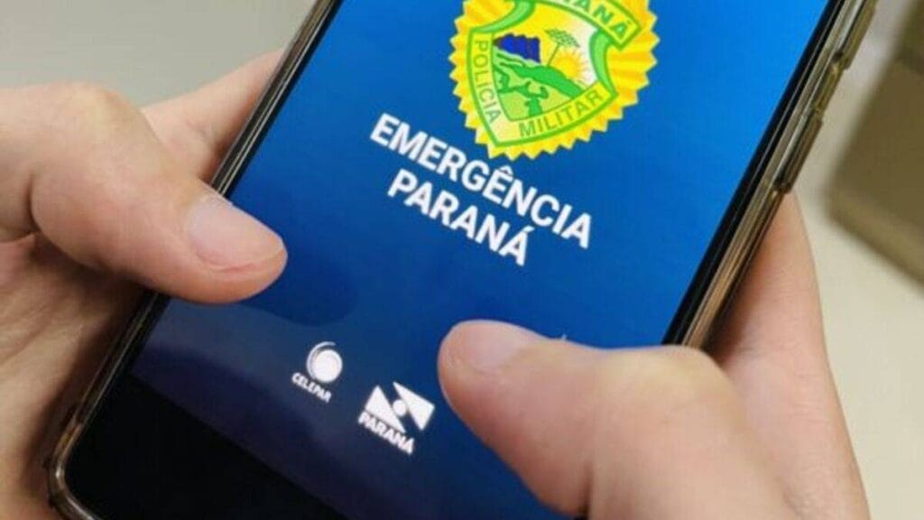 Pessoa segura um smartphone com a tela exibindo o aplicativo “Emergência Paraná”, da Polícia Militar do Paraná, com logotipos do governo e da Celepar.