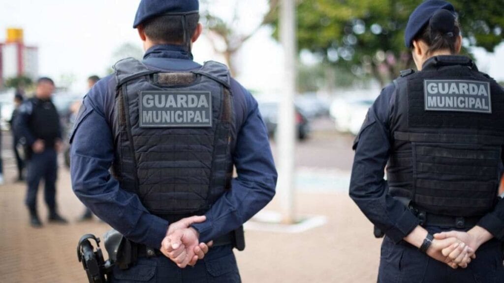 Dois agentes da Guarda Municipal - GCM, um homem e uma mulher, estão de costas, uniformizados e com coletes táticos, em ambiente urbano durante o dia.