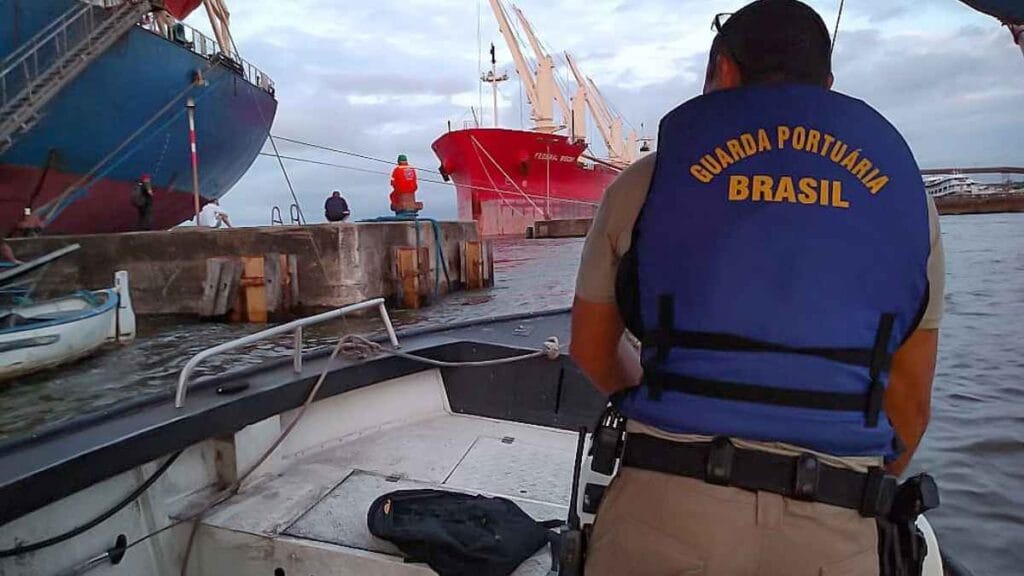Agente da Guarda Portuária do Brasil em embarcação, observando o cais onde há trabalhadores e navios cargueiros atracados. A cena ocorre ao entardecer, com destaque para o colete azul com inscrição “Guarda Portuária Brasil”.