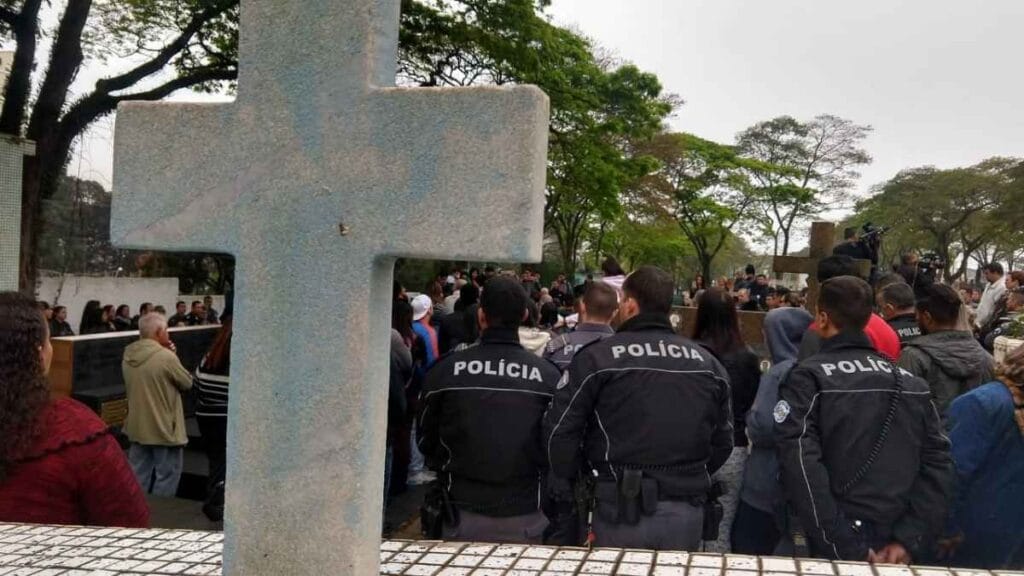 Policiais acompanham enterro de colega em cemitério de São Paulo, com cruz em destaque ao fundo.