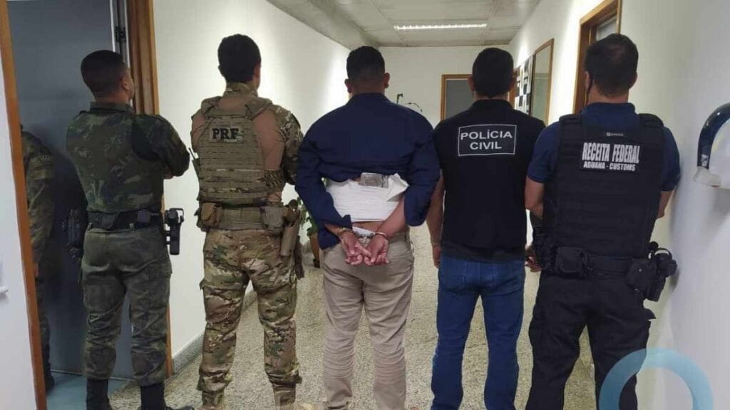 Equipes da Polícia Civil, PRF, Receita Federal e Exército escoltam um suspeito algemado com drogas presas ao corpo em corredor de unidade policial.