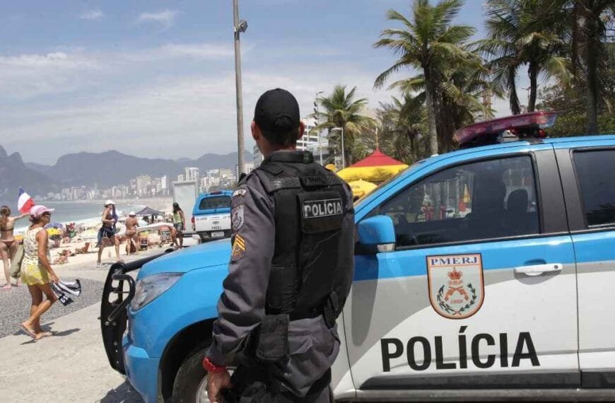 Policial militar da PMERJ patrulha área próxima à praia durante o dia, com viatura posicionada ao lado e banhistas ao fundo. A imagem mostra o policiamento ostensivo na orla do Rio de Janeiro, em cenário de grande movimento turístico e com presença de montanhas ao longe.
