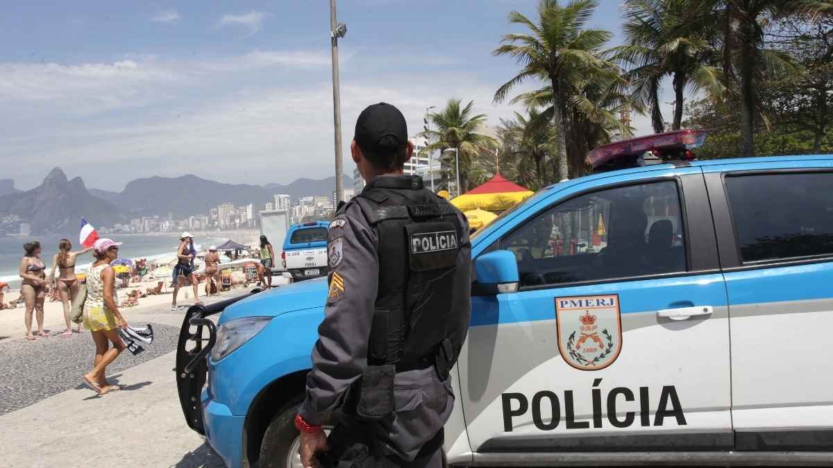 Policial militar da PMERJ patrulha área próxima à praia durante o dia, com viatura posicionada ao lado e banhistas ao fundo. A imagem mostra o policiamento ostensivo na orla do Rio de Janeiro, em cenário de grande movimento turístico e com presença de montanhas ao longe.