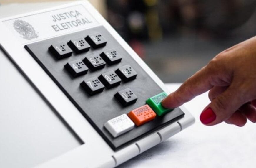 Pessoa votando em urna eletrônica da Justiça Eleitoral, com destaque para o dedo pressionando a tecla "CONFIRMA" na cor verde. A imagem mostra o teclado numérico com identificação em braile.