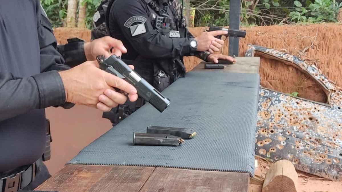 Porte de arma: Imagem mostra dois agentes armados em treinamento de tiro, manuseando pistolas sobre uma bancada com carregadores à frente. Ambos usam uniformes táticos pretos, sendo possível identificar o brasão da Polícia Penal Federal em um deles. Ao fundo, vê-se uma lataria de carro perfurada por disparos e vegetação densa. Cena típica de instrução prática voltada à segurança pública.