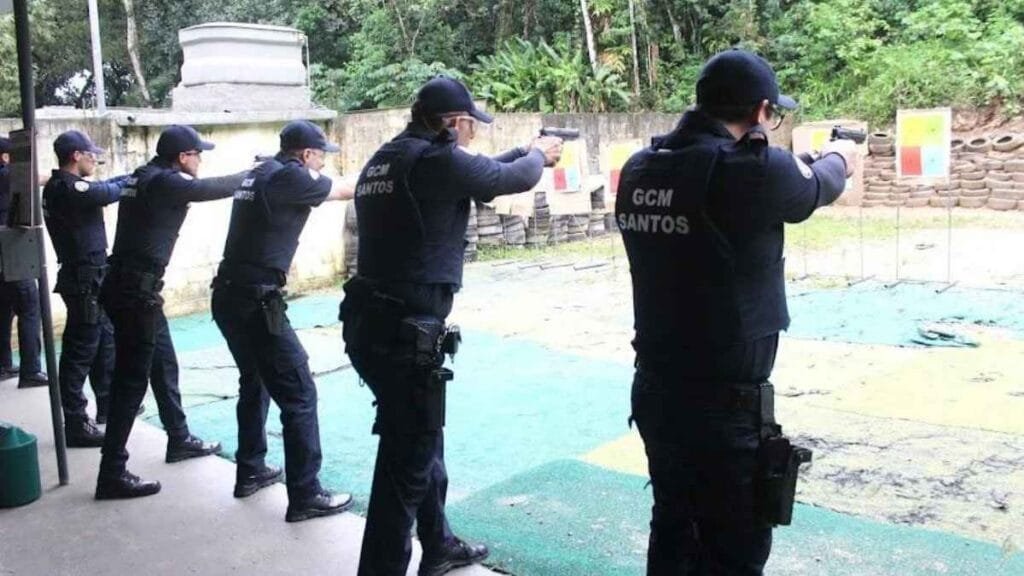 Porte de Arma: Agentes da Guarda Civil Municipal de Santos realizam treinamento de tiro em estande ao ar livre. Em posição de disparo, os guardas estão alinhados, usando uniforme tático com coletes identificados e armamento em punho. Ao fundo, há alvos coloridos dispostos em frente a barreiras de pneus e vegetação densa. A imagem destaca a capacitação e preparo da GCM para atuação em segurança pública.