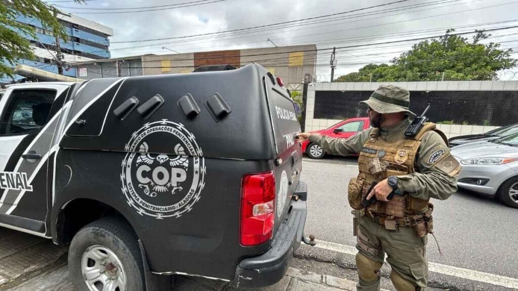 Recaptura: Policial penal fortemente equipado, com colete tático e rádio comunicador, inspeciona viatura do Comando de Operações Penitenciárias (COP) de Alagoas, estacionada em via urbana. Ao fundo, há carros e prédios comerciais.