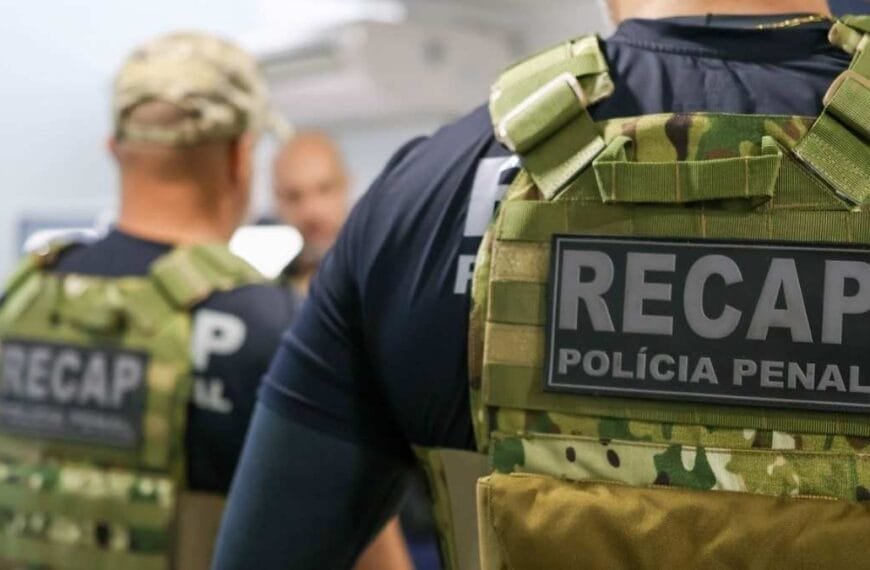Carreiras Policiais e desigualdade salarial no Brasil - Recaptura: Agentes da Polícia Penal com coletes camuflados identificados com a sigla "RECAP", em ambiente interno. O foco está nas costas de um dos agentes, evidenciando o emblema da equipe especializada em recaptura de foragidos.