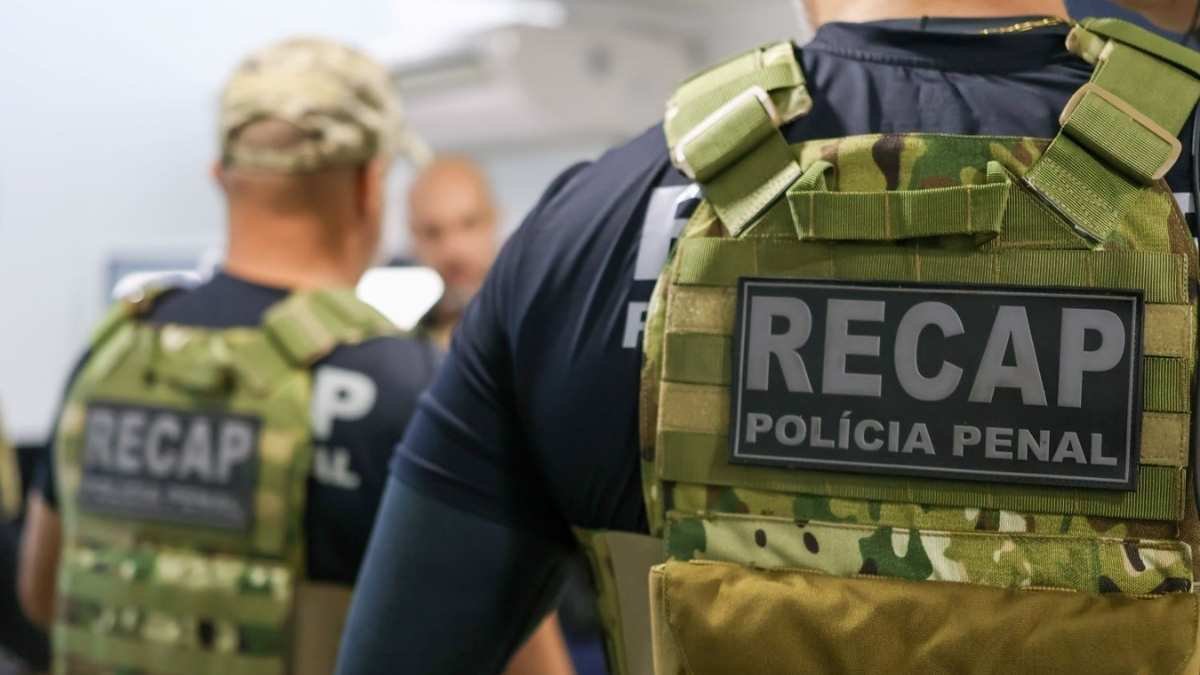 Carreiras Policiais e desigualdade salarial no Brasil - Recaptura: Agentes da Polícia Penal com coletes camuflados identificados com a sigla "RECAP", em ambiente interno. O foco está nas costas de um dos agentes, evidenciando o emblema da equipe especializada em recaptura de foragidos.