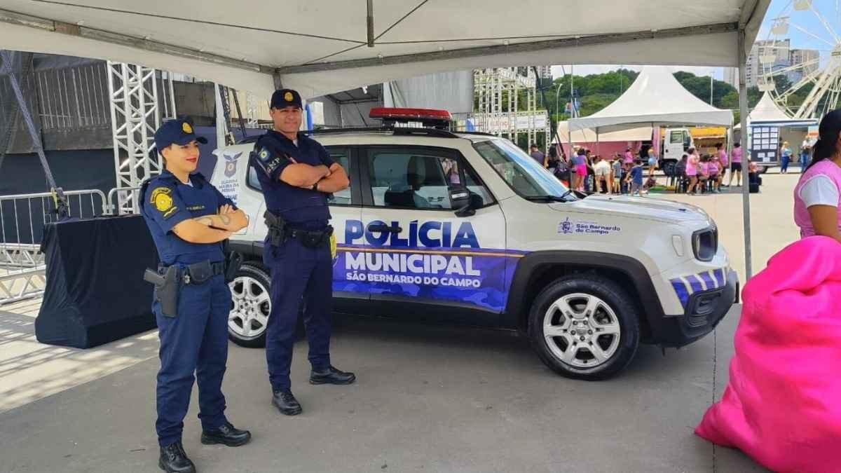 Polícia Municipal: Dois agentes uniformizados da Guarda Civil Municipal de São Bernardo do Campo posam ao lado de viatura caracterizada como "Polícia Municipal", durante evento público ao ar livre com tendas e público ao fundo.