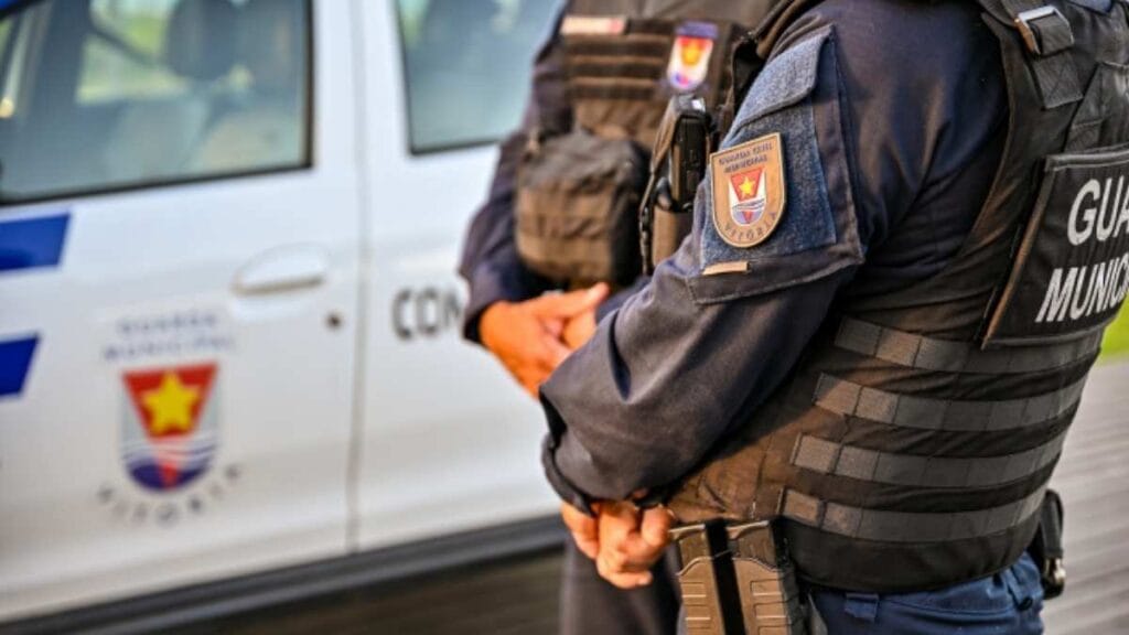 Dois agentes da Guarda Municipal de Vitória em frente a uma viatura oficial, usando uniformes táticos e coletes com identificação.