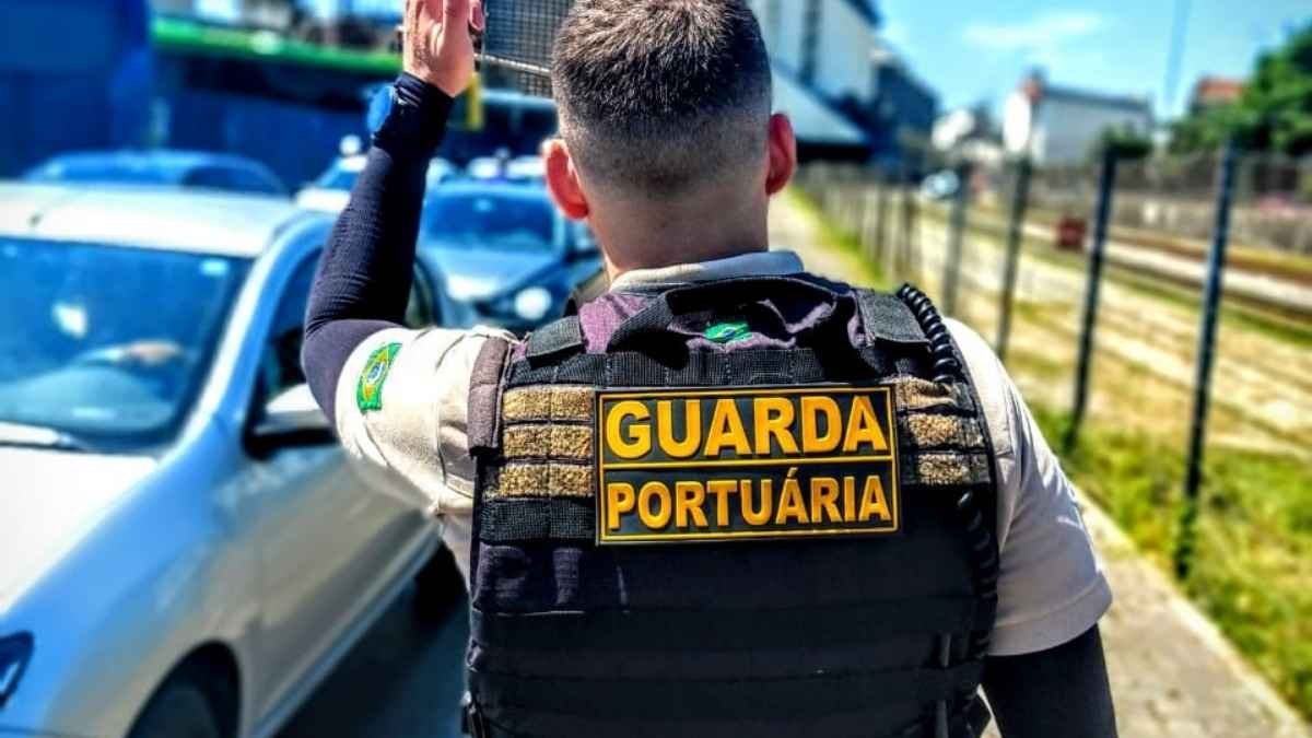 Agente da Guarda Portuária sinaliza o trânsito próximo a área portuária, usando colete tático com identificação visível nas costas. Ao fundo, veículos e estruturas típicas de zona portuária