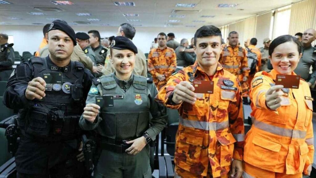 Policiais militares e bombeiros do Ceará posam sorridentes segurando cartões, durante cerimônia em ambiente fechado, vestindo uniformes operacionais e boinas