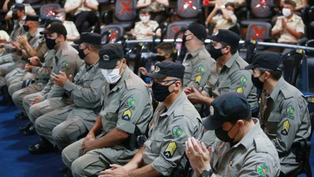 Serviço voluntário: Policiais militares de Goiás, uniformizados e com máscaras, participam de solenidade em ambiente fechado, sentados e aplaudindo.