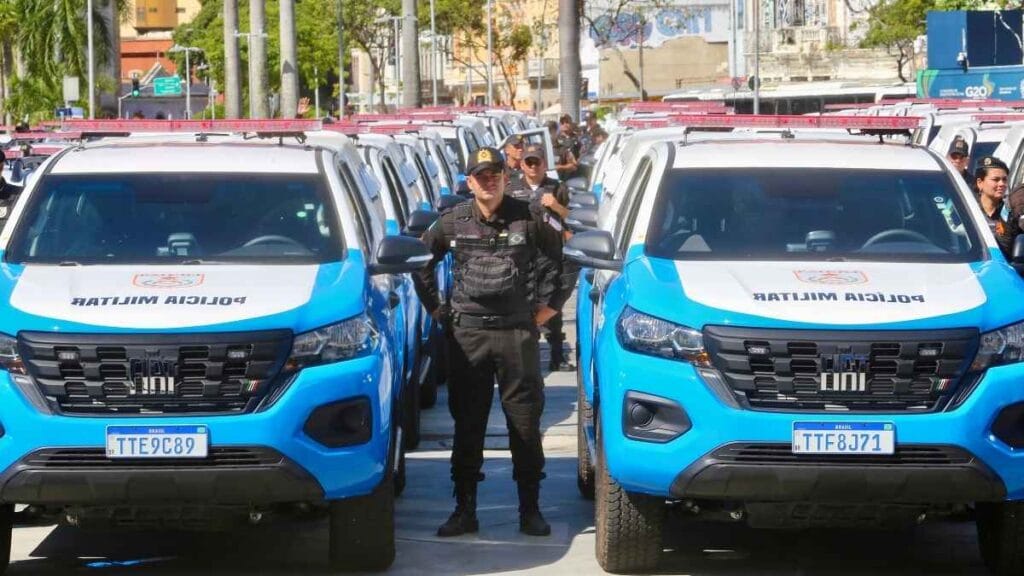 Policiais militares do Rio de Janeiro posam entre dezenas de novas viaturas da PMERJ alinhadas em pátio aberto, durante cerimônia oficial de entrega dos veículos.