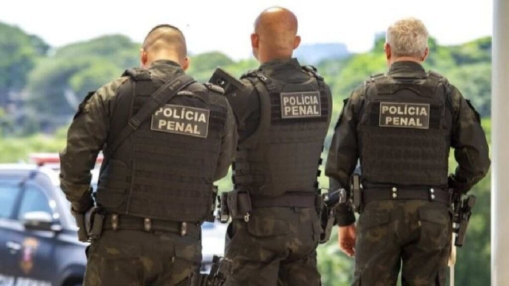 Três agentes da Polícia Penal de uniforme tático vistos de costas durante operação ou patrulha.