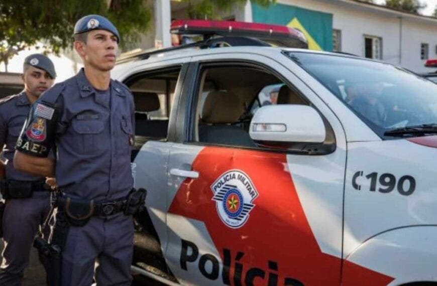 2029: qualquer cargo na PM exigirá NS: Policiais militares do BAEP, da Polícia Militar de São Paulo, estão posicionados ao lado de uma viatura oficial com as portas abertas. Os agentes estão fardados e atentos, com o prédio da corporação e a bandeira do Brasil ao fundo, em um cenário de prontidão e disciplina.