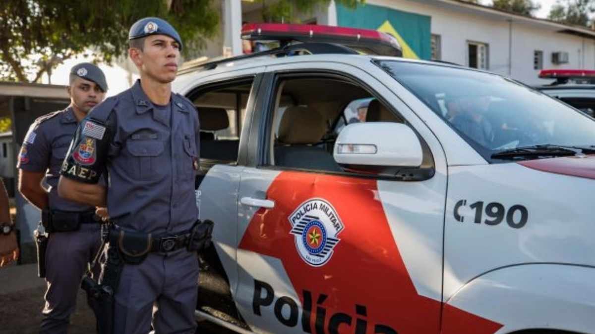 2029: qualquer cargo na PM exigirá NS: Policiais militares do BAEP, da Polícia Militar de São Paulo, estão posicionados ao lado de uma viatura oficial com as portas abertas. Os agentes estão fardados e atentos, com o prédio da corporação e a bandeira do Brasil ao fundo, em um cenário de prontidão e disciplina.
