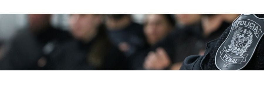 **Texto ALT resumido:**
Insígnia da Polícia Penal de Santa Catarina em destaque, com agentes desfocados ao fundo.
