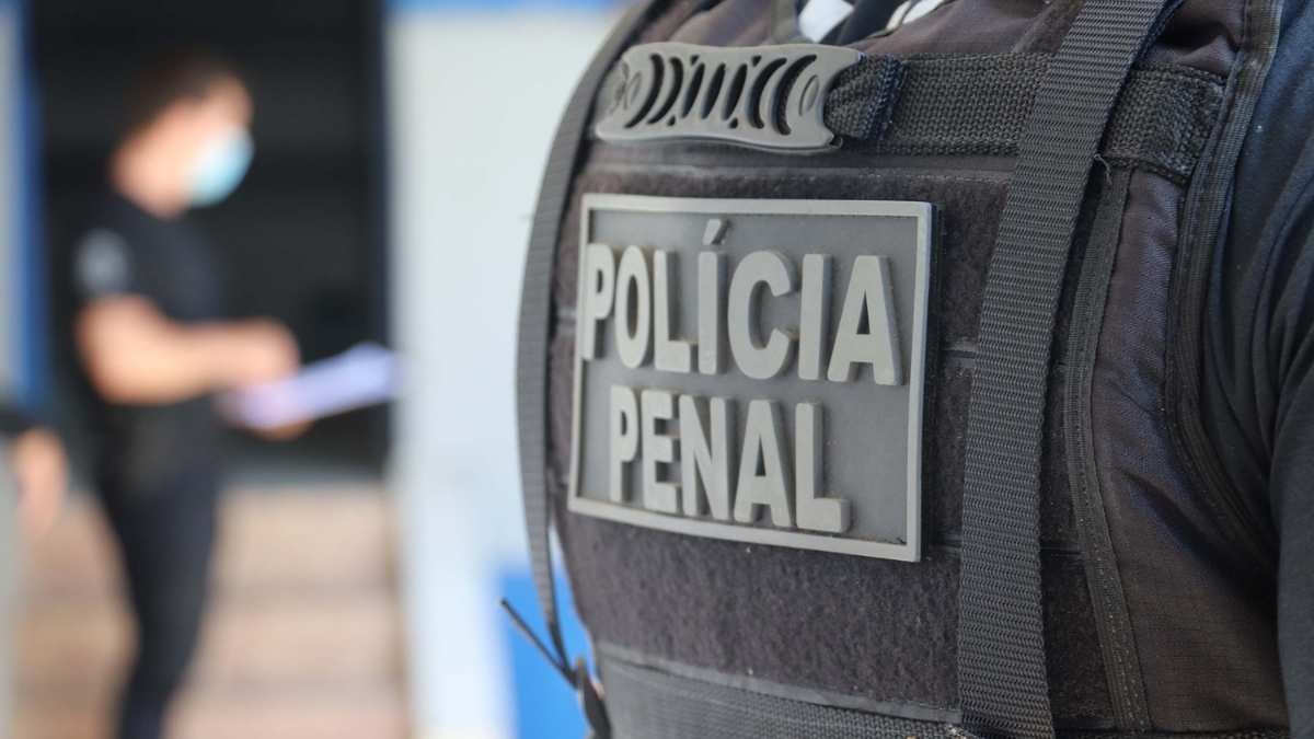 Colete tático com identificação "Polícia Penal" em destaque, usado por agente de segurança; ao fundo, pessoa desfocada com máscara facial segura documentos.