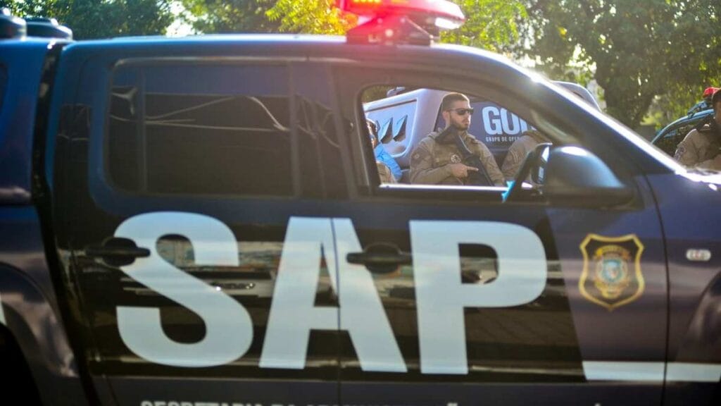 Viatura caracterizada da Secretaria da Administração Penitenciária do Ceará (SAP) em destaque, com sirene acionada e agentes uniformizados visíveis no interior do veículo. Ao fundo, é possível ver outro carro com a inscrição "GOP" (Grupo de Operações Penitenciárias), sugerindo a atuação em operação ou evento oficial.