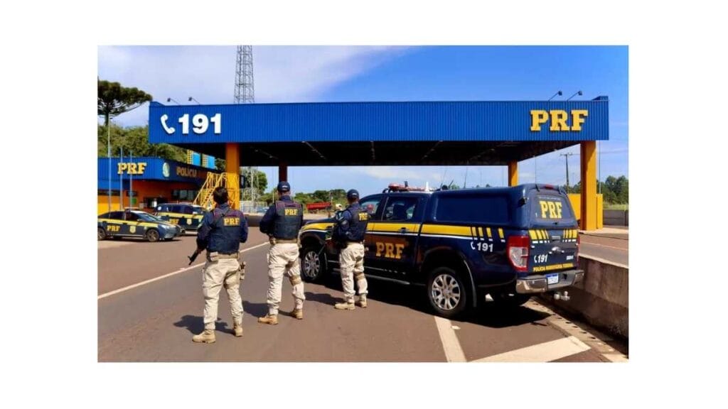 Policiais da PRF (Polícia Rodoviária Federal) uniformizados ao lado de viaturas na frente de um posto policial com estrutura azul e amarela, com a sigla “PRF” e o número de emergência “191” visíveis.