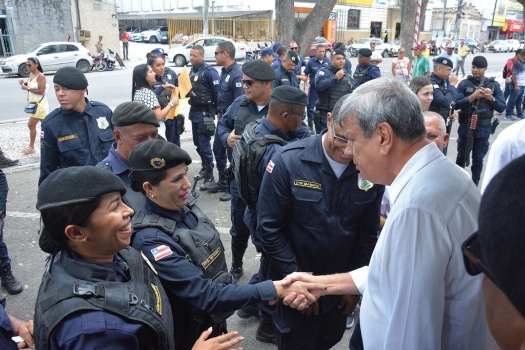 Na imagem, um grupo de guardas municipais uniformizados está reunido em uma rua, interagindo com um homem de camisa branca, que aparenta ser uma autoridade. Ele cumprimenta calorosamente uma das agentes, enquanto outras pessoas observam e registram o momento. O ambiente é descontraído e demonstra integração entre a autoridade e os agentes de segurança pública. Ao fundo, há veículos estacionados e mais pessoas, sugerindo que o encontro ocorre em um espaço urbano e público.