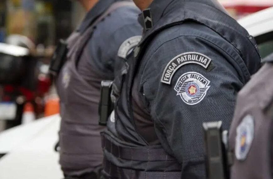 Efetivo: Policiais militares de São Paulo, com uniformes da "Força Patrulha", estão perfilados em ação ostensiva nas ruas da cidade. O foco da imagem está no brasão da "Polícia Militar do Estado de São Paulo" presente na farda. Ao fundo, há trânsito urbano e cones de sinalização, sugerindo operação policial em andamento.
