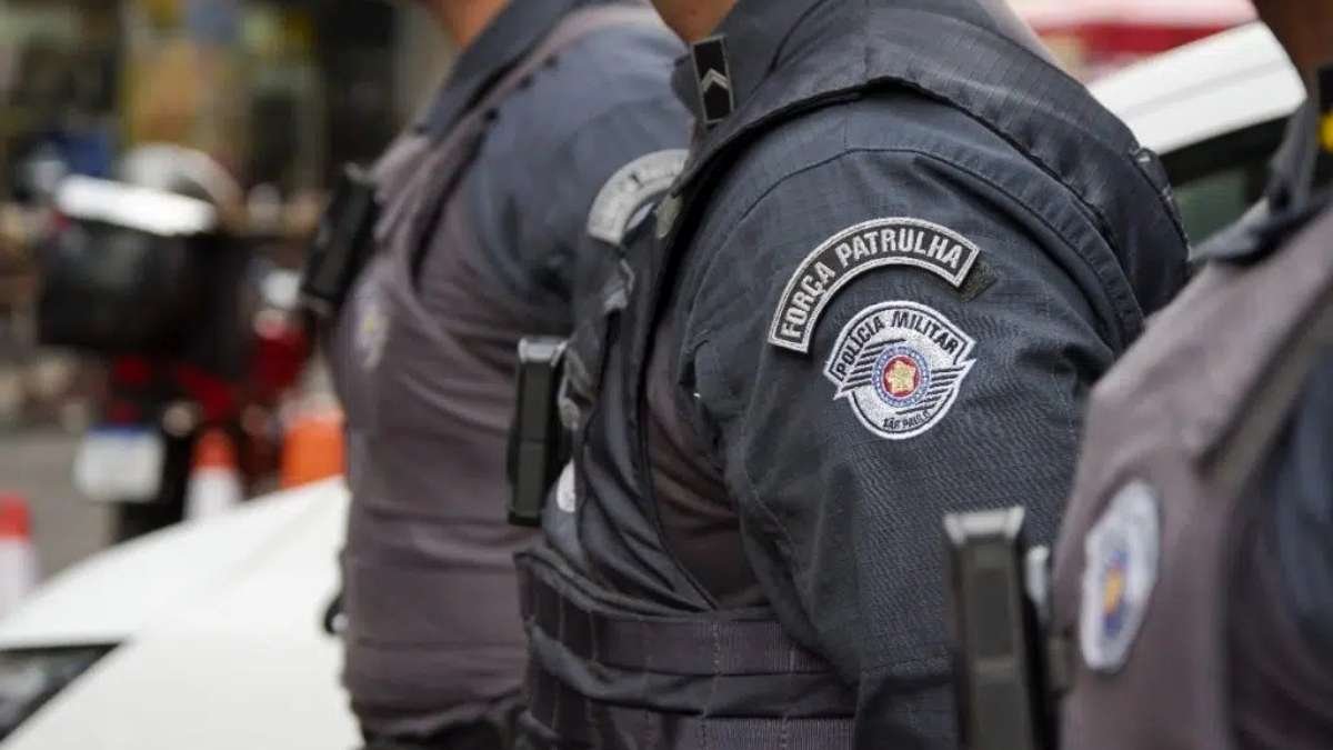 Efetivo: Policiais militares de São Paulo, com uniformes da "Força Patrulha", estão perfilados em ação ostensiva nas ruas da cidade. O foco da imagem está no brasão da "Polícia Militar do Estado de São Paulo" presente na farda. Ao fundo, há trânsito urbano e cones de sinalização, sugerindo operação policial em andamento.