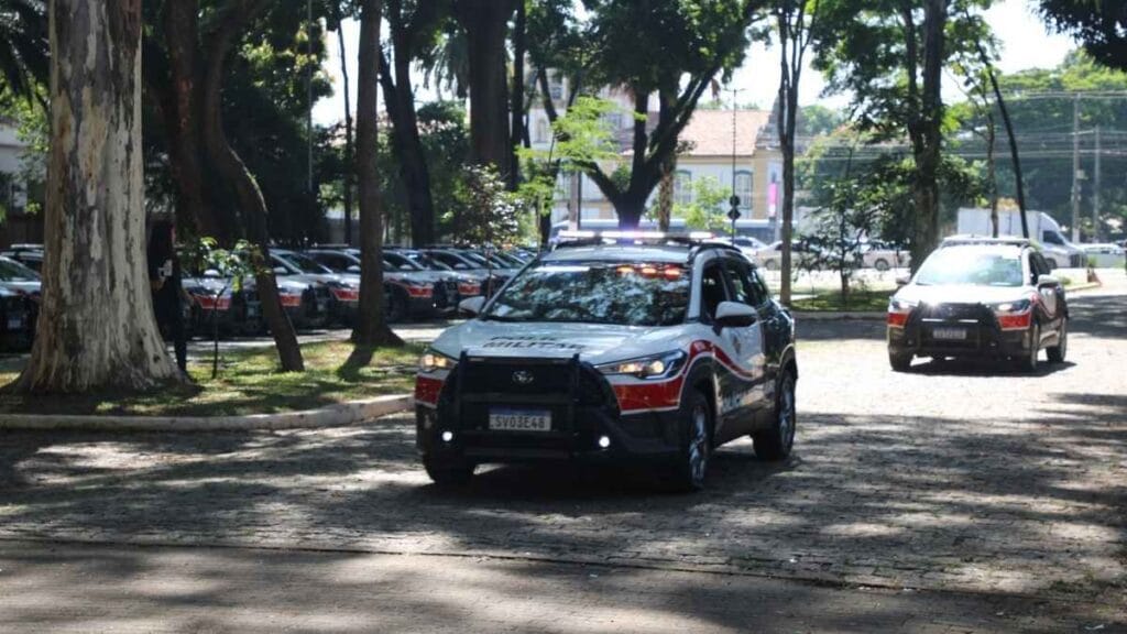 SUVs: Viaturas da Polícia Militar de São Paulo em movimento dentro de área arborizada; à frente, SUV caracterizado com pintura oficial e giroflex acionado, enquanto outras viaturas permanecem estacionadas ao fundo.