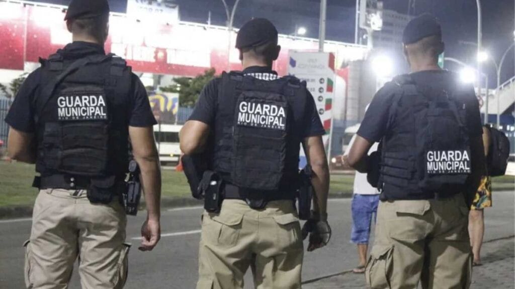 Três agentes da Guarda Municipal do Rio de Janeiro, de uniforme e colete tático, caminham em patrulhamento noturno em área urbana.