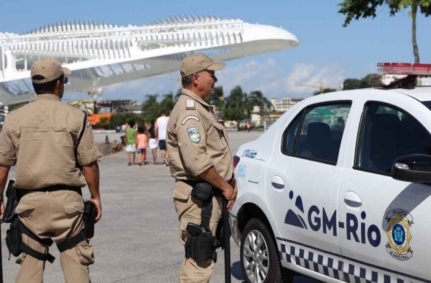 ADPFs questionam Força Municipal do Rio: Dois agentes da Guarda Municipal do Rio de Janeiro estão em patrulhamento próximo a uma viatura da corporação, em área urbana com pedestres ao fundo.