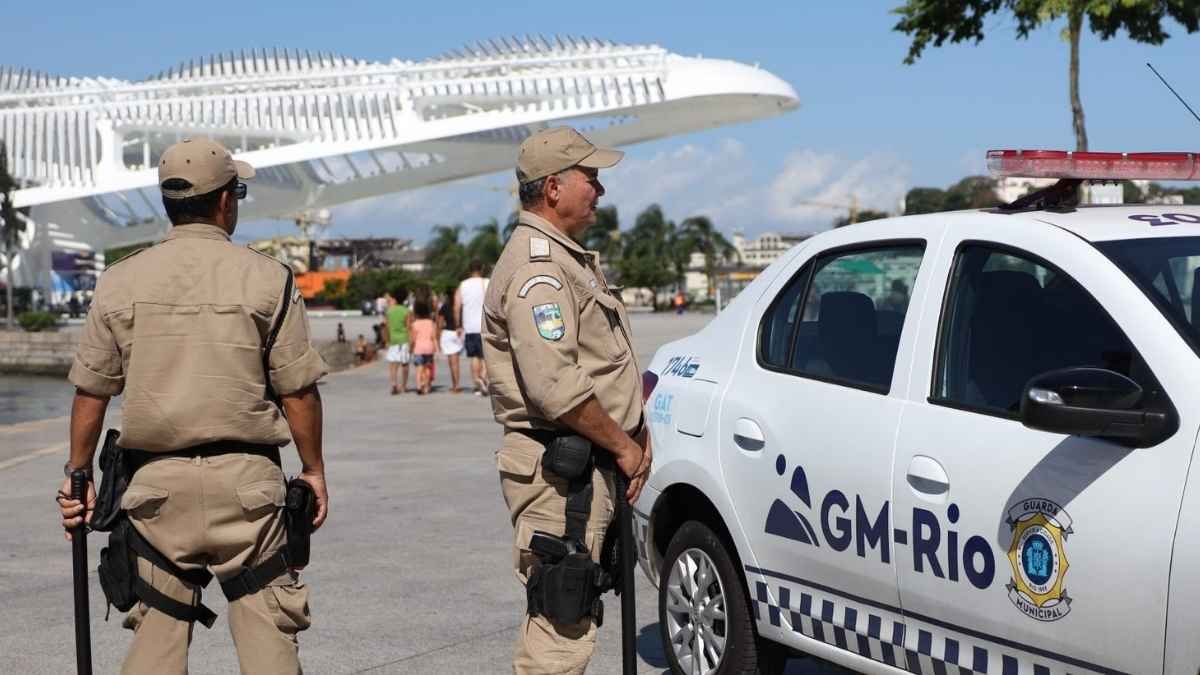 ADPFs questionam Força Municipal do Rio: Dois agentes da Guarda Municipal do Rio de Janeiro estão em patrulhamento próximo a uma viatura da corporação, em área urbana com pedestres ao fundo.