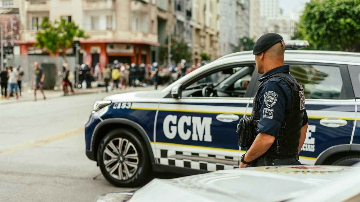 Agente da Guarda Civil Metropolitana de São Paulo em pé ao lado de viatura da corporação durante operação em via urbana.
