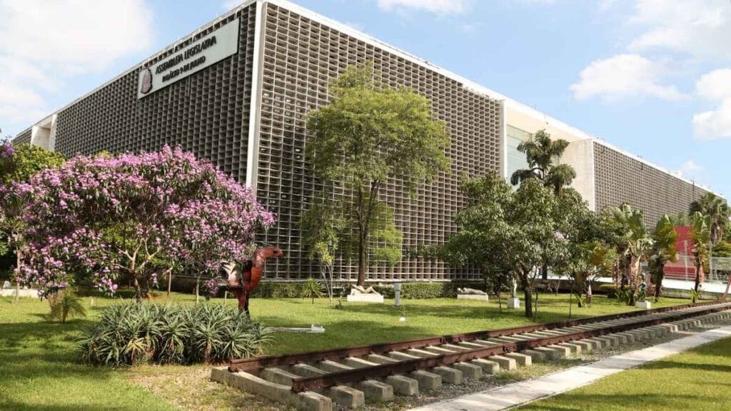 Lei Orgânica da PCSP: **Texto ALT resumido:** Fachada da Assembleia Legislativa do Estado de São Paulo (ALESP), com jardim arborizado e árvores floridas em primeiro plano.