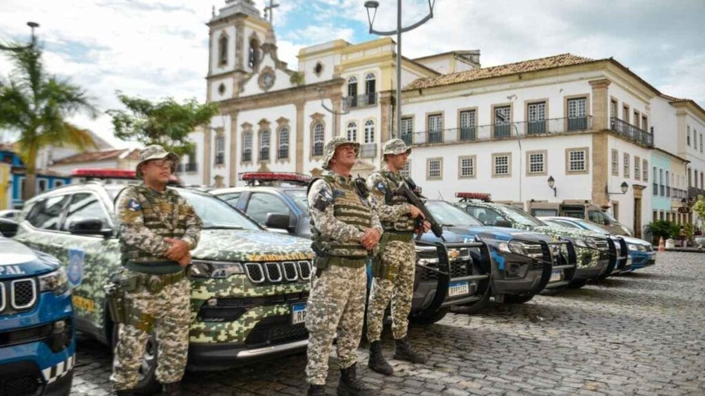 Os grupos especiais da GCM de Salvador: Três agentes da Guarda Civil Municipal de Salvador, fardados com uniforme camuflado em tons de verde, posam em frente a uma fileira de viaturas oficiais estacionadas em via de paralelepípedos. Ao fundo, construções históricas e uma igreja colonial compõem o cenário.