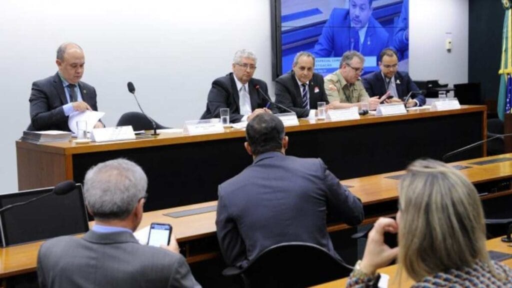 Ciclo completo de polícia: Audiência pública na Câmara dos Deputados com cinco homens sentados à mesa, incluindo um oficial militar, discutindo tema relacionado à segurança pública. Participantes no plenário acompanham a sessão, enquanto um telão ao fundo exibe a imagem ampliada de um dos presentes.