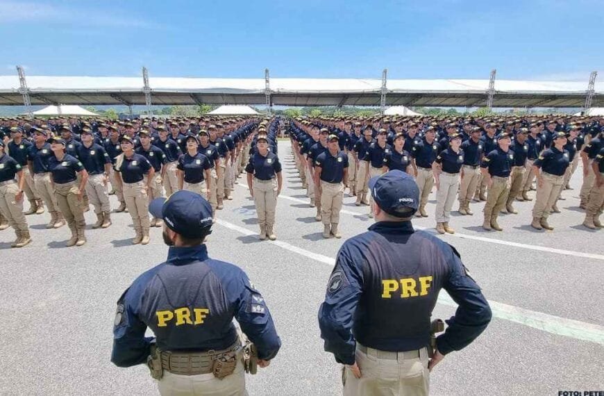 Ciclo completo de polícia: A imagem mostra um grande contingente de policiais da PRF enfileirados em formação, usando uniformes azul-marinho e calças bege, com dois policiais de costas, em primeiro plano, supervisionando a tropa.