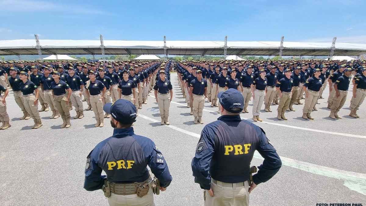 Ciclo completo de polícia: A imagem mostra um grande contingente de policiais da PRF enfileirados em formação, usando uniformes azul-marinho e calças bege, com dois policiais de costas, em primeiro plano, supervisionando a tropa.