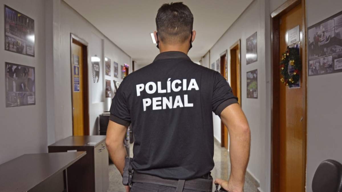 Agente da Polícia Penal caminha por corredor interno, vestindo uniforme preto com os dizeres 'POLÍCIA PENAL' nas costas. PPTO: chefias renunciam em massa