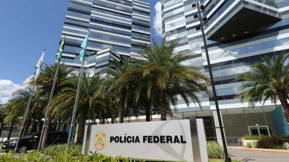 Polícia Federal: grande demais? A imagem mostra a fachada da sede da Polícia Federal, com letreiro e brasão da instituição em primeiro plano, bandeiras hasteadas e prédios modernos ao fundo, cercados por palmeiras.