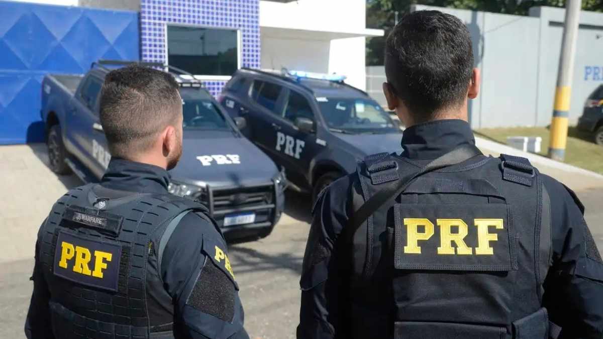Aposentadoria Policial: Dois policiais da PRF de costas observam viaturas estacionadas em frente a uma unidade da corporação.