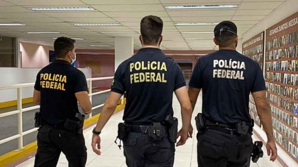 A imagem mostra três policiais federais de costas, caminhando por um corredor interno. Eles vestem camisetas pretas com a inscrição “Polícia Federal” em letras amarelas e portam equipamentos na cintura.