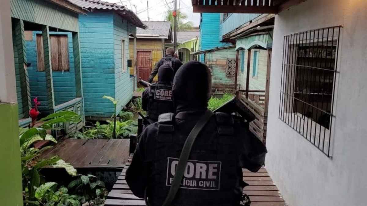Domínio das fações: Policiais civis do CORE, armados e usando uniformes táticos, caminham por passarelas de madeira entre casas simples de madeira colorida em área de palafitas, durante operação.