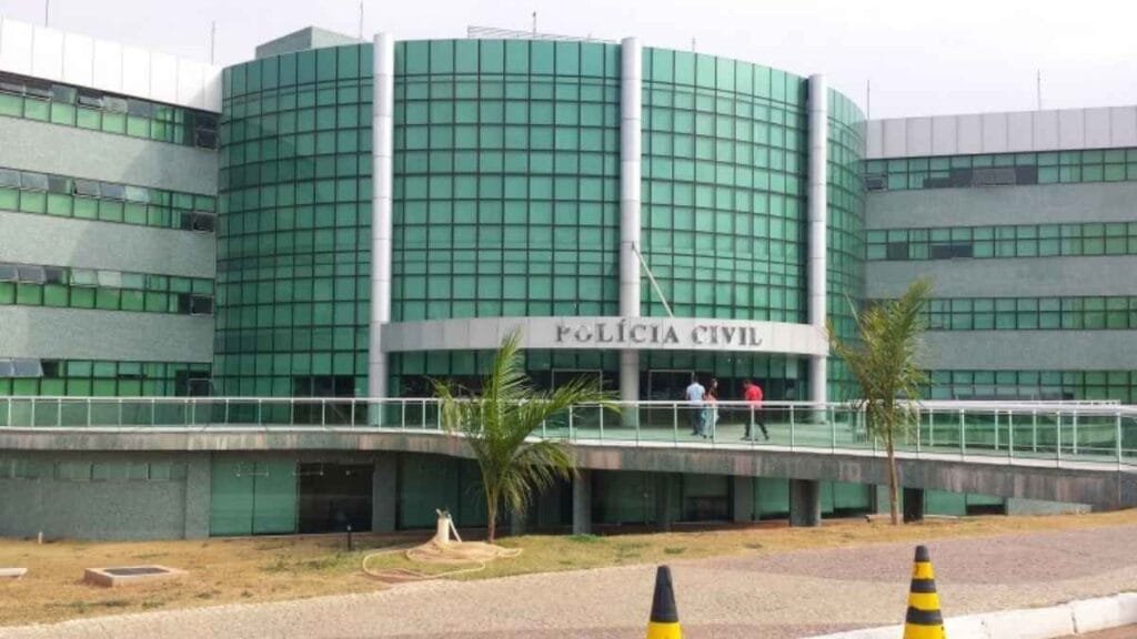 Equiparação PCDF e PF: Fachada do prédio da Polícia Civil do Distrito Federal (PCDF), com arquitetura moderna e vidros verdes. Três pessoas caminham sobre a passarela de acesso ao edifício.