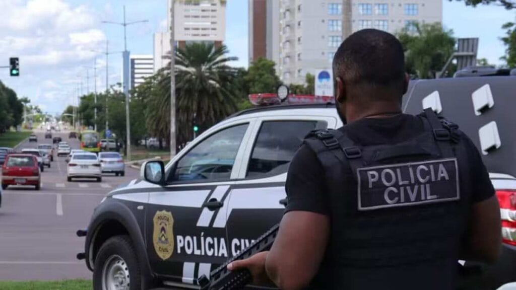 Unificação dos cargos de escrivão e agente: Policial civil armado observa o trânsito em via urbana, ao lado de viatura caracterizada da Polícia Civil, durante operação ou patrulhamento diurno.