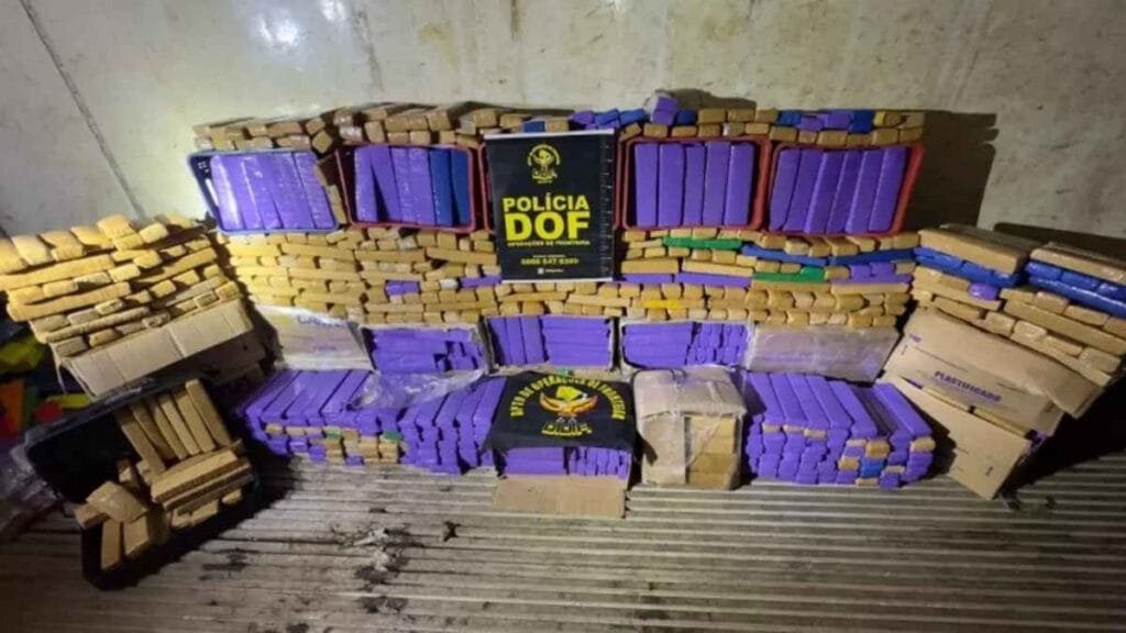 O Brasil como entreposto das frogas: Pilhas de tabletes de drogas apreendidas, embalados em cores roxa, amarela e azul, empilhados junto a caixas de papelão. No centro, placa do DOF (Departamento de Operações de Fronteira) da Polícia Militar de Mato Grosso do Sul.